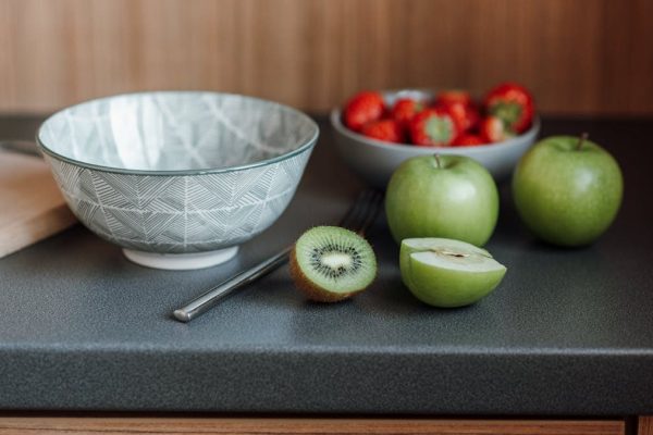 Table with bowl near fork and green apples with kiwi and cutting board near strawberries in dishware in light kitchen